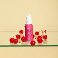 Lubrificante Liio Dafne a base d’olio, flacone su superficie trasparente con ciliegie, sfondo chiaro