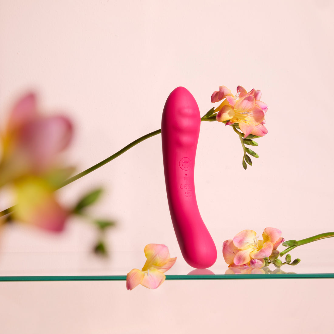 Vibratore rosso Double Trouble su superficie trasparente, con fiori e sfondo rosa tenue